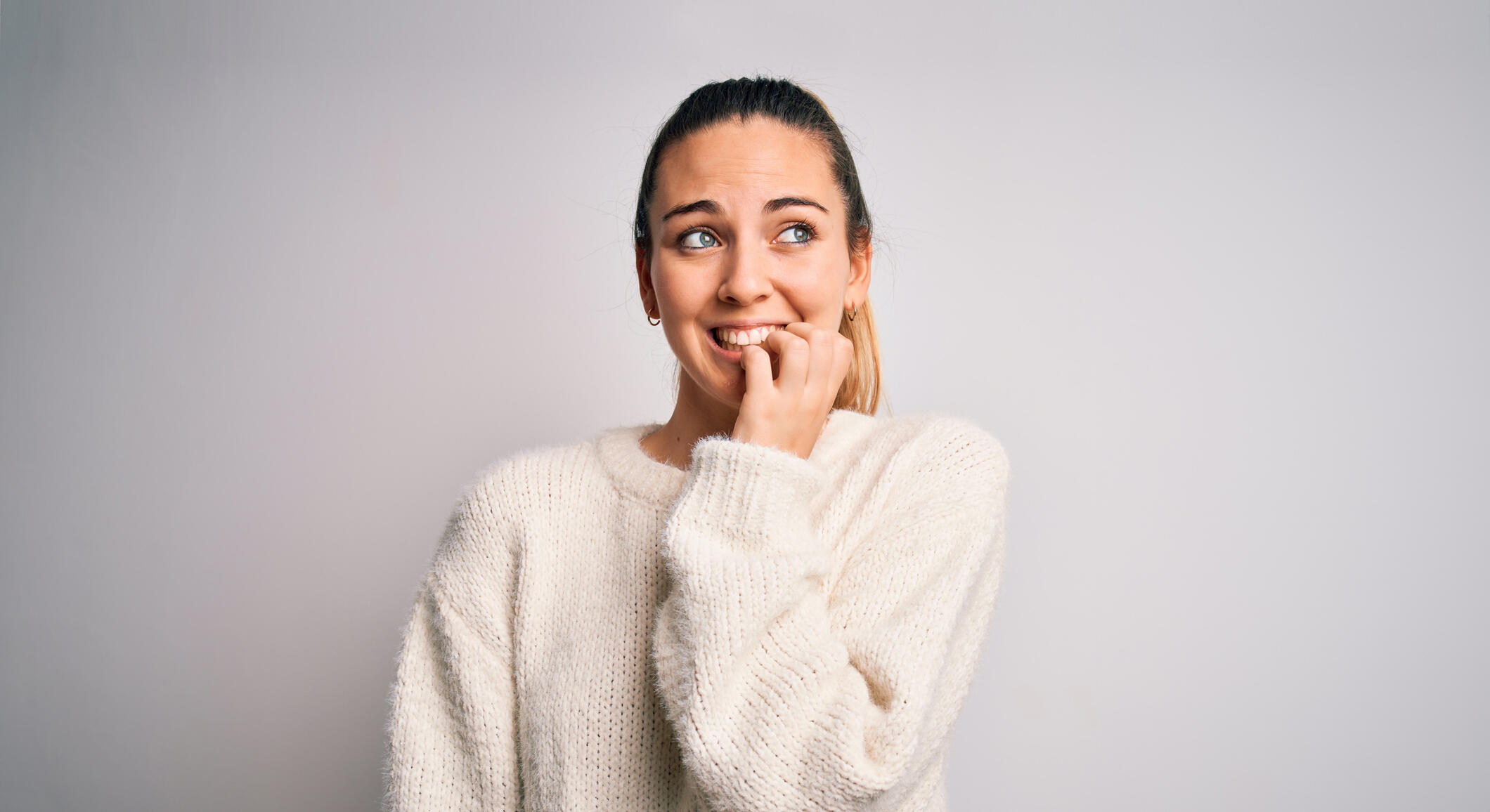 Young beautiful blonde woman with blue eyes wearing casual sweater over white background looking stressed and nervous with hands on mouth biting nails. Anxiety problem.